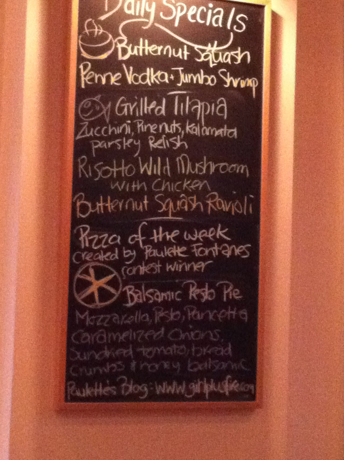 Poorly lit restaurant chalkboard listing daily specials, including butternut squash penne vodka + jumbo shrimp and pizza of the week created by Paulette Fontanes (sic) Balsamic Pesto Pie