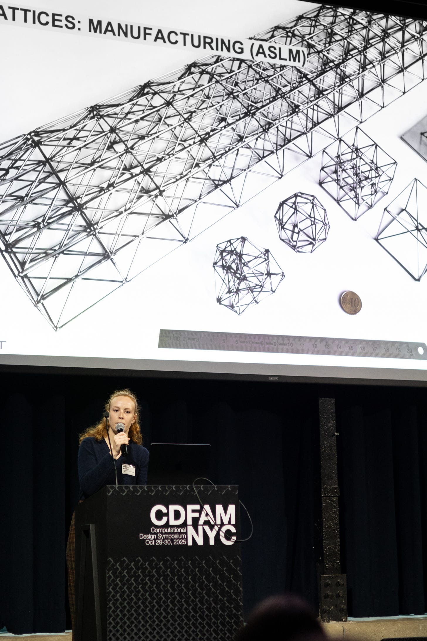 A speaker presenting at CDFAM NYC 2025, with a projected slide showing an abstract design featuring lattice structures and various geometric shapes in a scientific context.