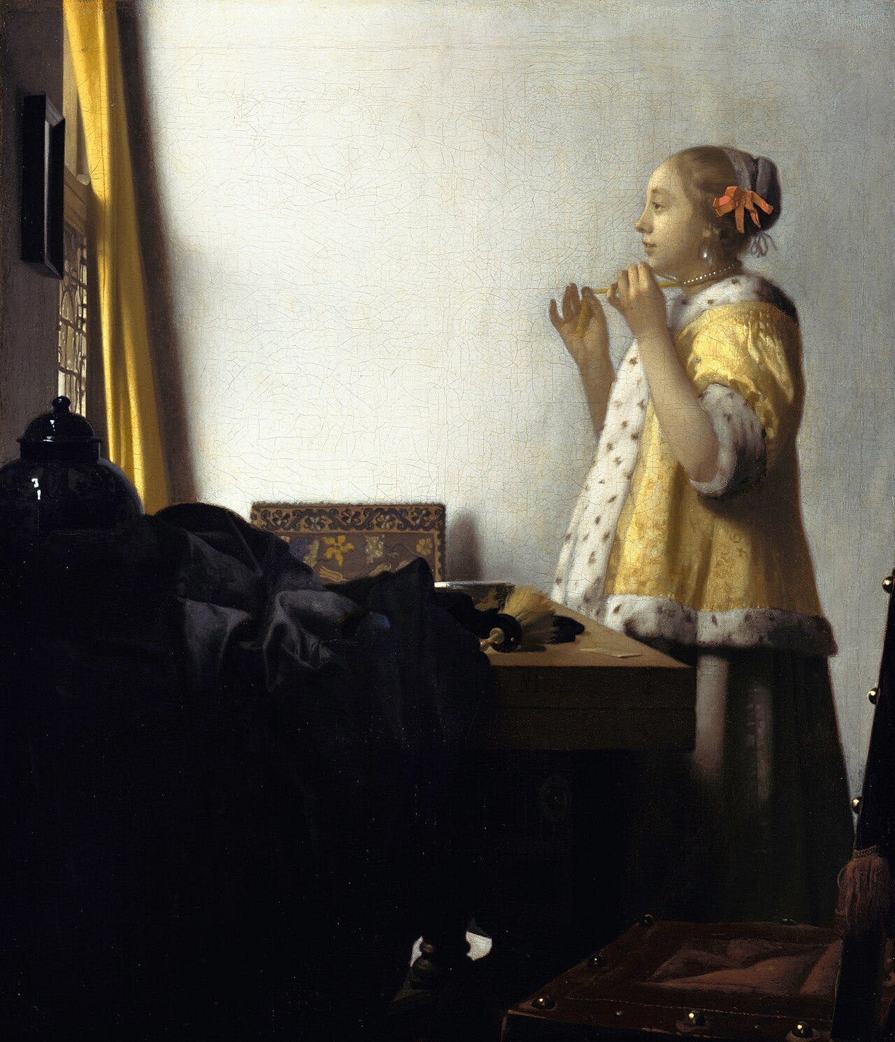 Young woman in a yellow fur trimmed jacket looking into a mirror while tying a pearl necklace Young woman in a yellow fur trimmed jacket looking into a mirror while tying a pearl necklace