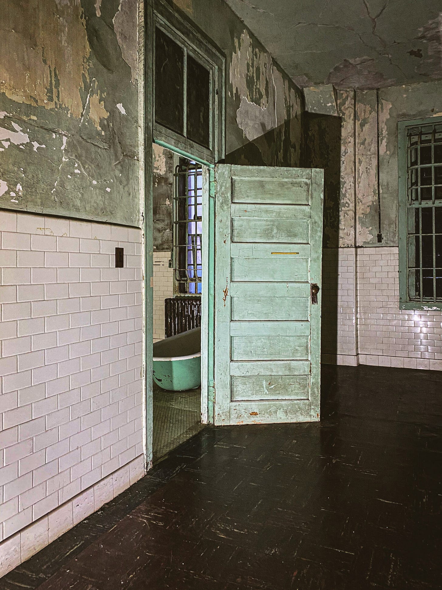 The interior of a derelict building with dark floors and walls of white tile and peeling green paint. An open doorway reveals a glimpse of a mint green bathtub in an adjacent room. The interior of a derelict building with dark floors and walls of white tile and peeling green paint. An open doorway reveals a glimpse of a mint green bathtub in an adjacent room.