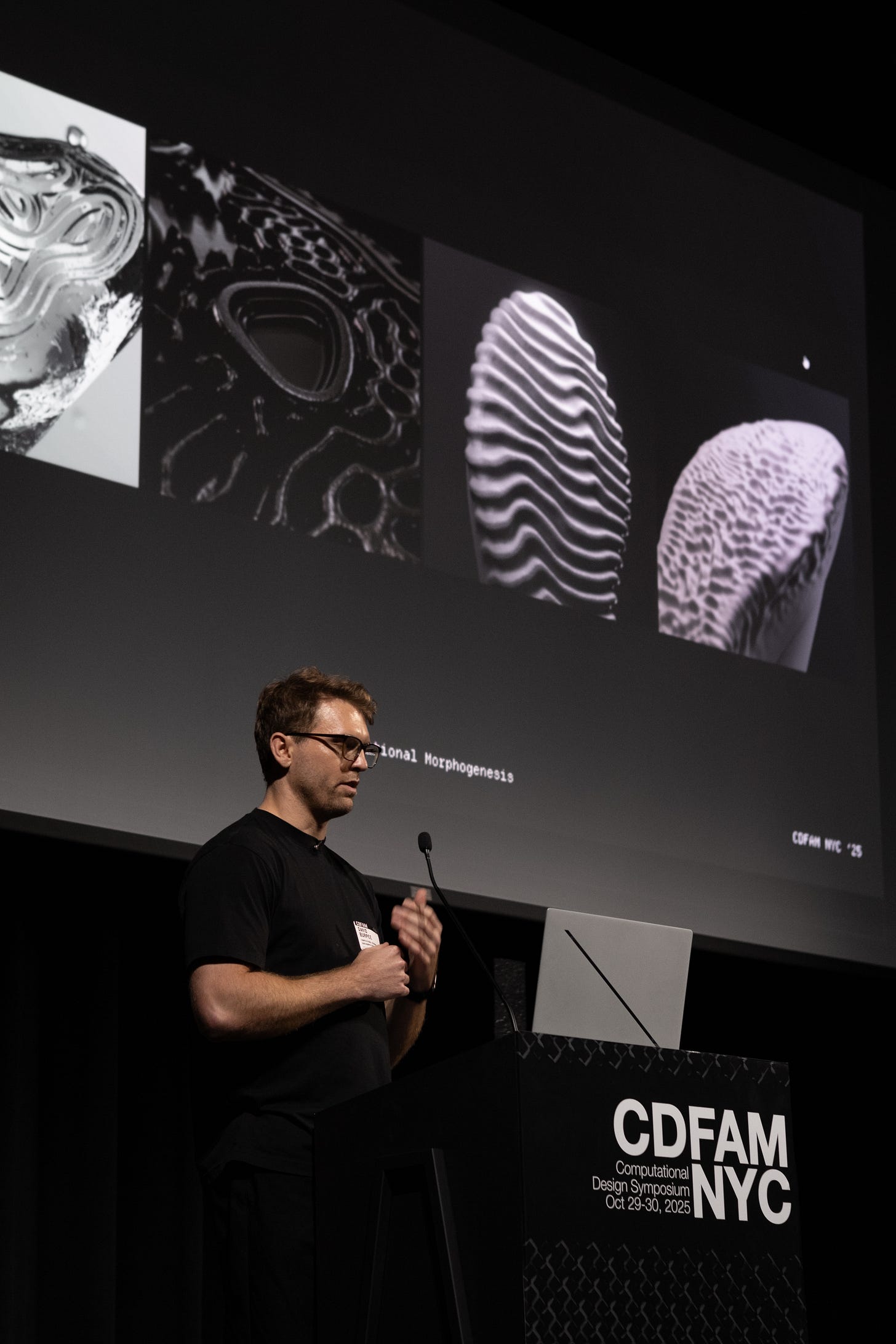 A presenter speaking at a conference about computational design, with abstract visuals displayed on a screen behind. The visuals include various morphogenetic patterns and textures.