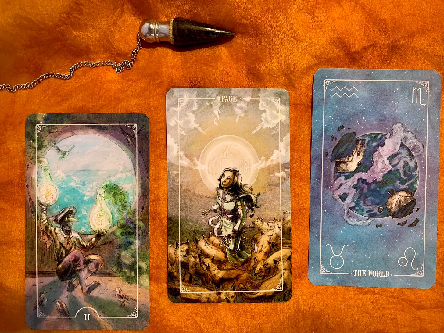 Three tarot cards and a pendulum on an orange silk background. The cards, from left to right, are the two of coins (a man balancing on one foot while holding alchemical bottles in each hand), the page of coins (depicting a young girl on a mountaintop surrounded by a sea of foxes at her feet), and the World (in which an image of the earth floats in space, with large portions of the land breaking off as it forms and reforms itself).