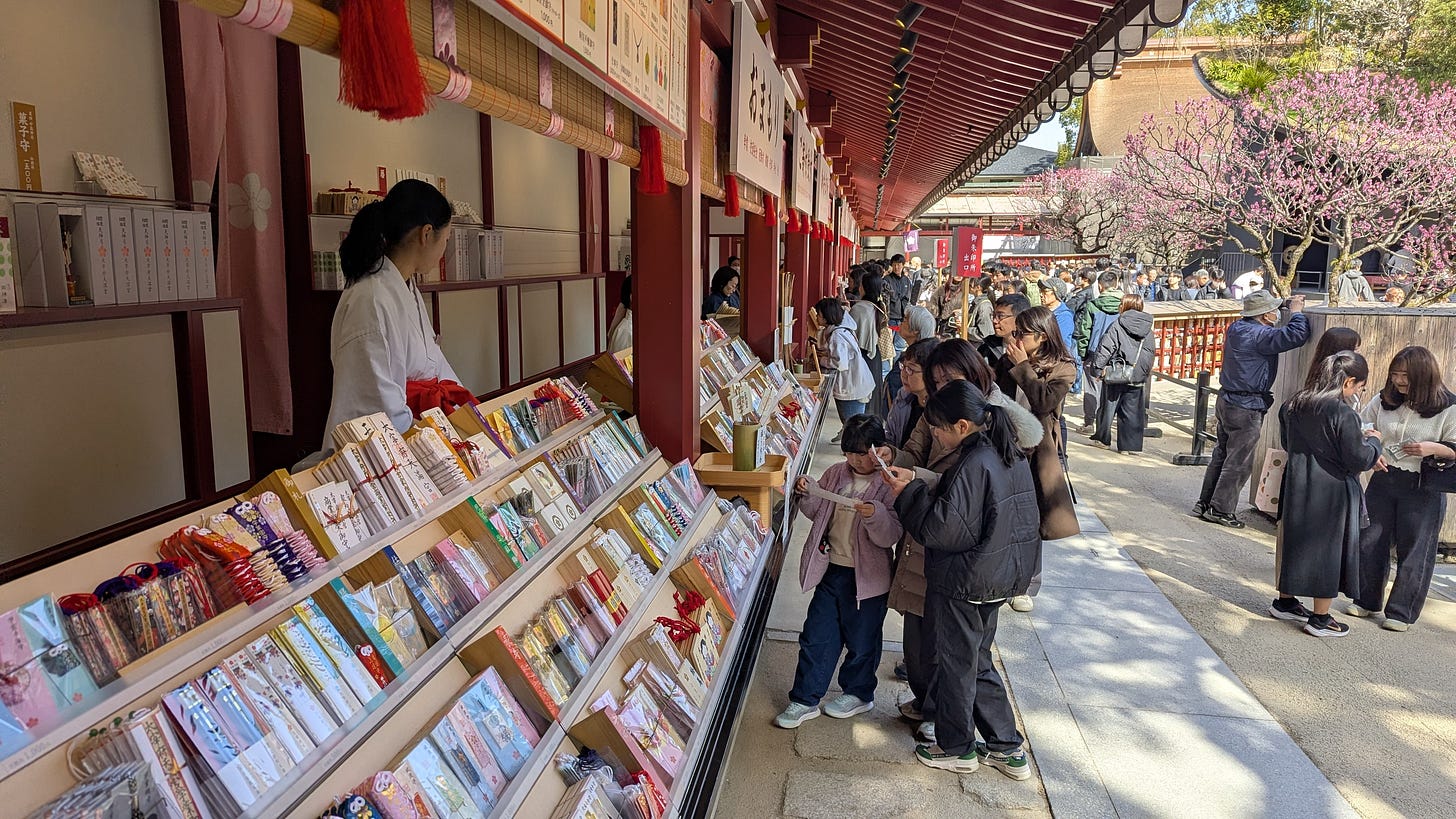 Children choose an omamori (lucky amulet) from rows of colorful choices while a shrine maiden looks on.