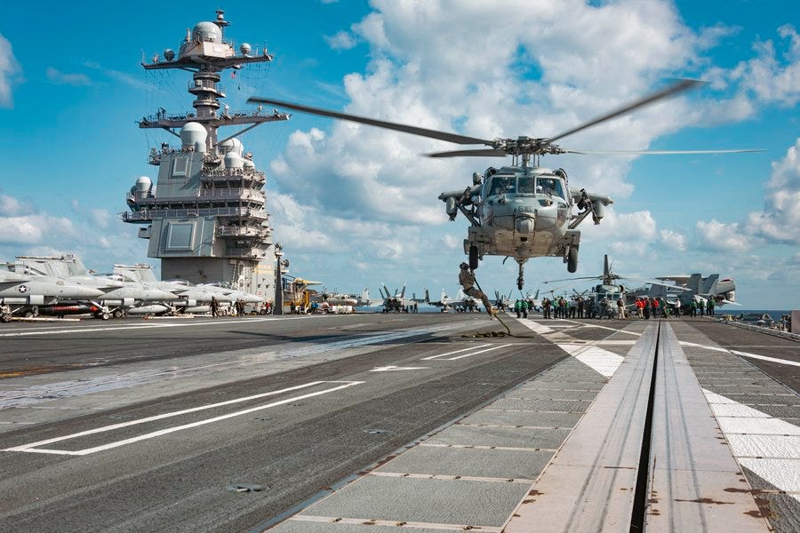 The image captures a dynamic scene on the flight deck of the USS Gerald R. Ford (CVN-78), where sailors from Explosive Ordnance Disposal Mobile Unit (EODMU) 12 are fast roping from an MH-60S Sea Hawk helicopter. The helicopter is prominently positioned in the center of the image, hovering above the deck with its rotors spinning, creating a sense of motion and urgency. The aircraft carrier's deck is visible, showcasing various military aircraft and equipment, emphasizing the operational environment. The backdrop includes the towering structure of the carrier, with clear skies and some clouds adding to the dramatic setting. This image highlights the readiness and capabilities of the U.S. Navy, focusing on the specialized training and operations of the EOD team. The image captures a dynamic scene on the flight deck of the USS Gerald R. Ford (CVN-78), where sailors from Explosive Ordnance Disposal Mobile Unit (EODMU) 12 are fast roping from an MH-60S Sea Hawk helicopter. The helicopter is prominently positioned in the center of the image, hovering above the deck with its rotors spinning, creating a sense of motion and urgency. The aircraft carrier's deck is visible, showcasing various military aircraft and equipment, emphasizing the operational environment. The backdrop includes the towering structure of the carrier, with clear skies and some clouds adding to the dramatic setting. This image highlights the readiness and capabilities of the U.S. Navy, focusing on the specialized training and operations of the EOD team.