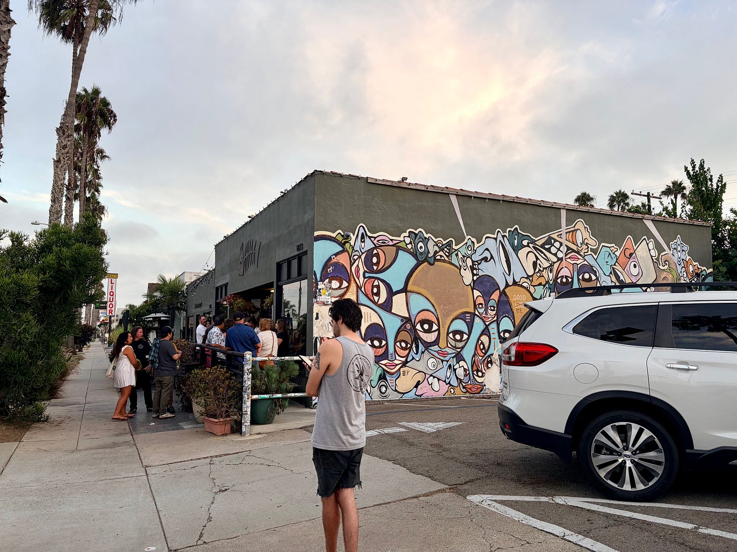 A crowd gathers on the sidewalk outside Wrench and Rodent Seabasstropub in the evening, beneath a colorful mural of stylized faces, with palm trees lining the street and a car parked nearby.