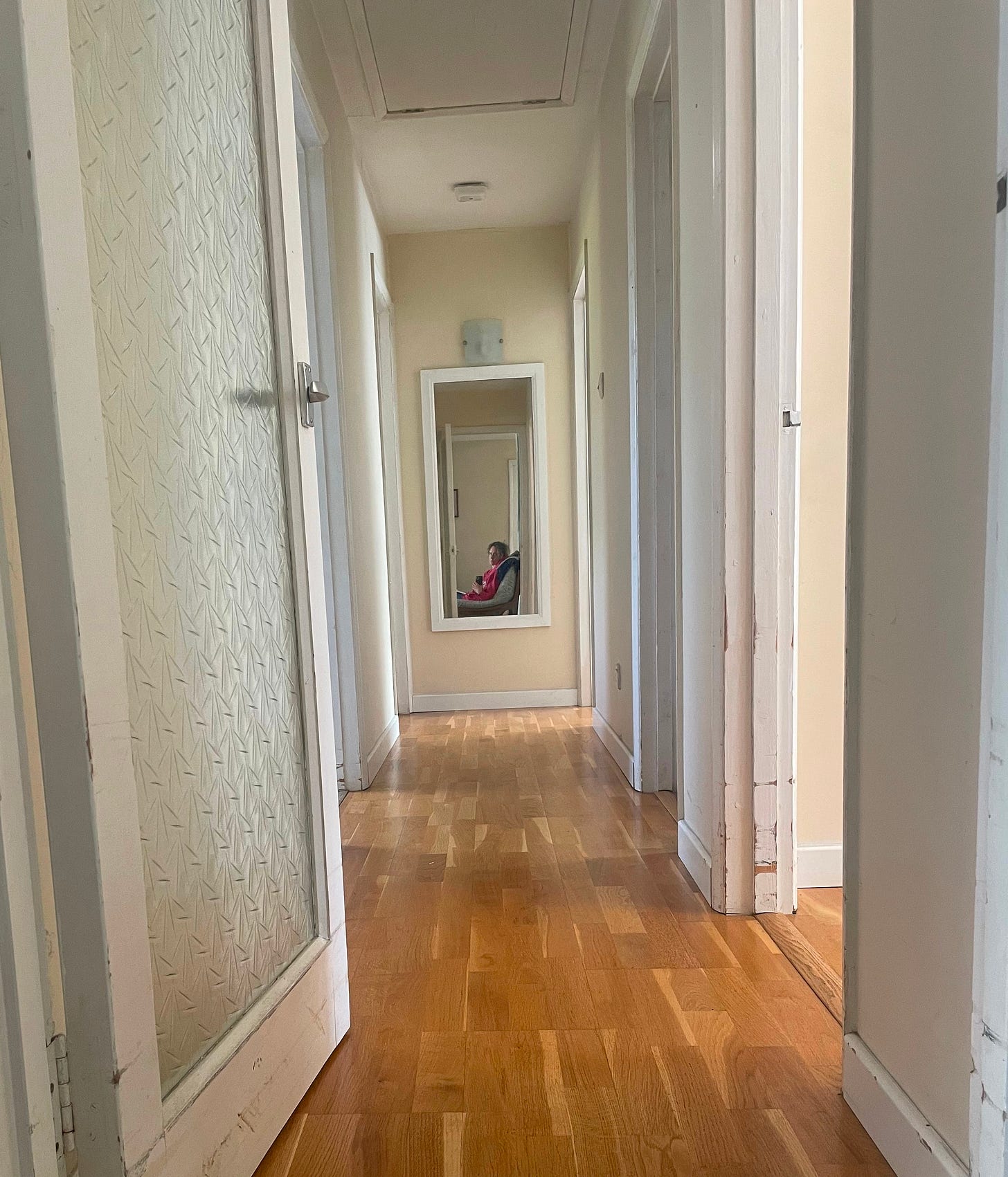 A corridor with doors on either side, ending in a mirror which shows the subject taking the photo - a woman wearing a pink jumper A corridor with doors on either side, ending in a mirror which shows the subject taking the photo - a woman wearing a pink jumper