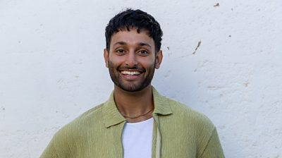 Jas wears a white t-shirt under a green shirt with a gold chain 