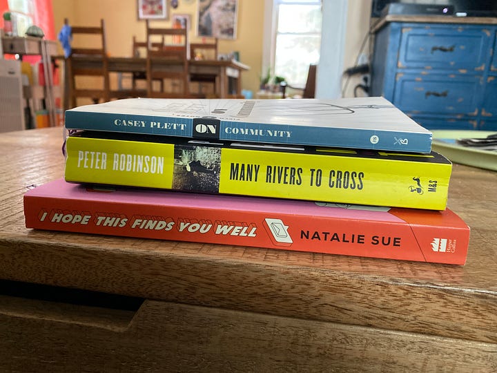 In one photo, we see in the foreground a stack of three books, On Community, Many Rivers to Cross, and I Hope This Finds You Well. In the other photo, we see the cover image of the audiobook The Mistress of Bhatia House, where an illustration shows a woman with a long, pink headscarf looking out to the water.