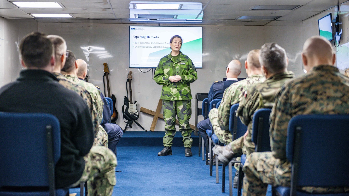 A Swedish brigadier general in camouflage uniform stands in front of a presentation screen addressing rows of seated officers, almost all of them men. A Swedish brigadier general in camouflage uniform stands in front of a presentation screen addressing rows of seated officers, almost all of them men.