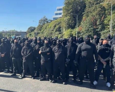 A large group of men dressed in black, many of them wearing balaclavas and other face coverings, block a road A large group of men dressed in black, many of them wearing balaclavas and other face coverings, block a road