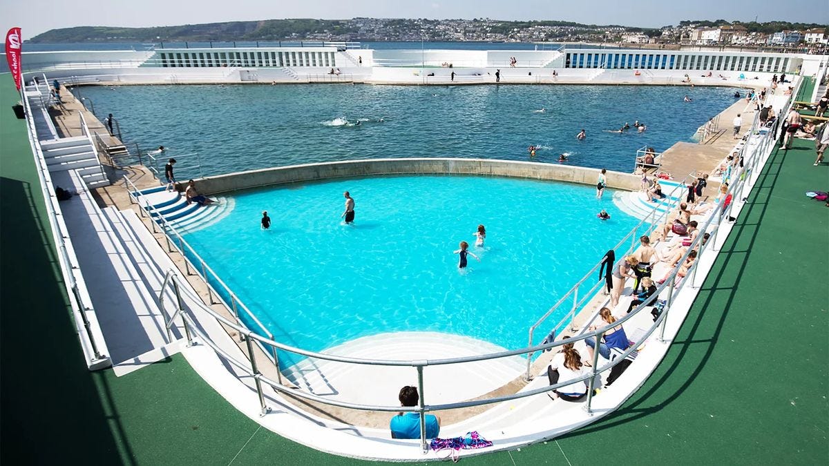 Outdoor lido with turquoise and saltwater pools, swimmers relaxing under clear blue sky beside the coast.