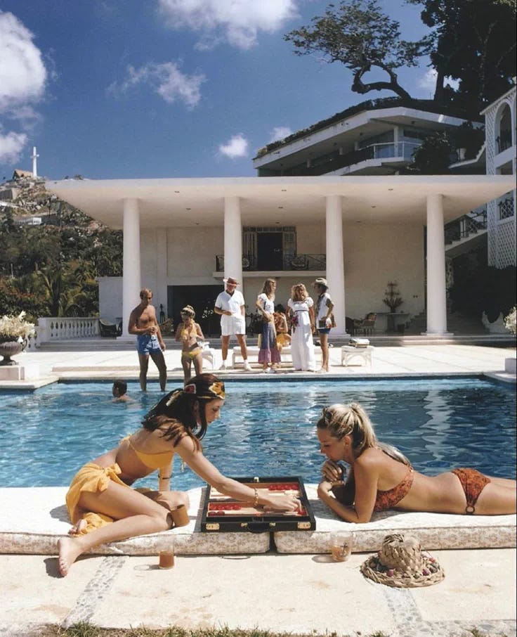 This may contain: two women sitting on the edge of a swimming pool in front of a large white house This may contain: two women sitting on the edge of a swimming pool in front of a large white house