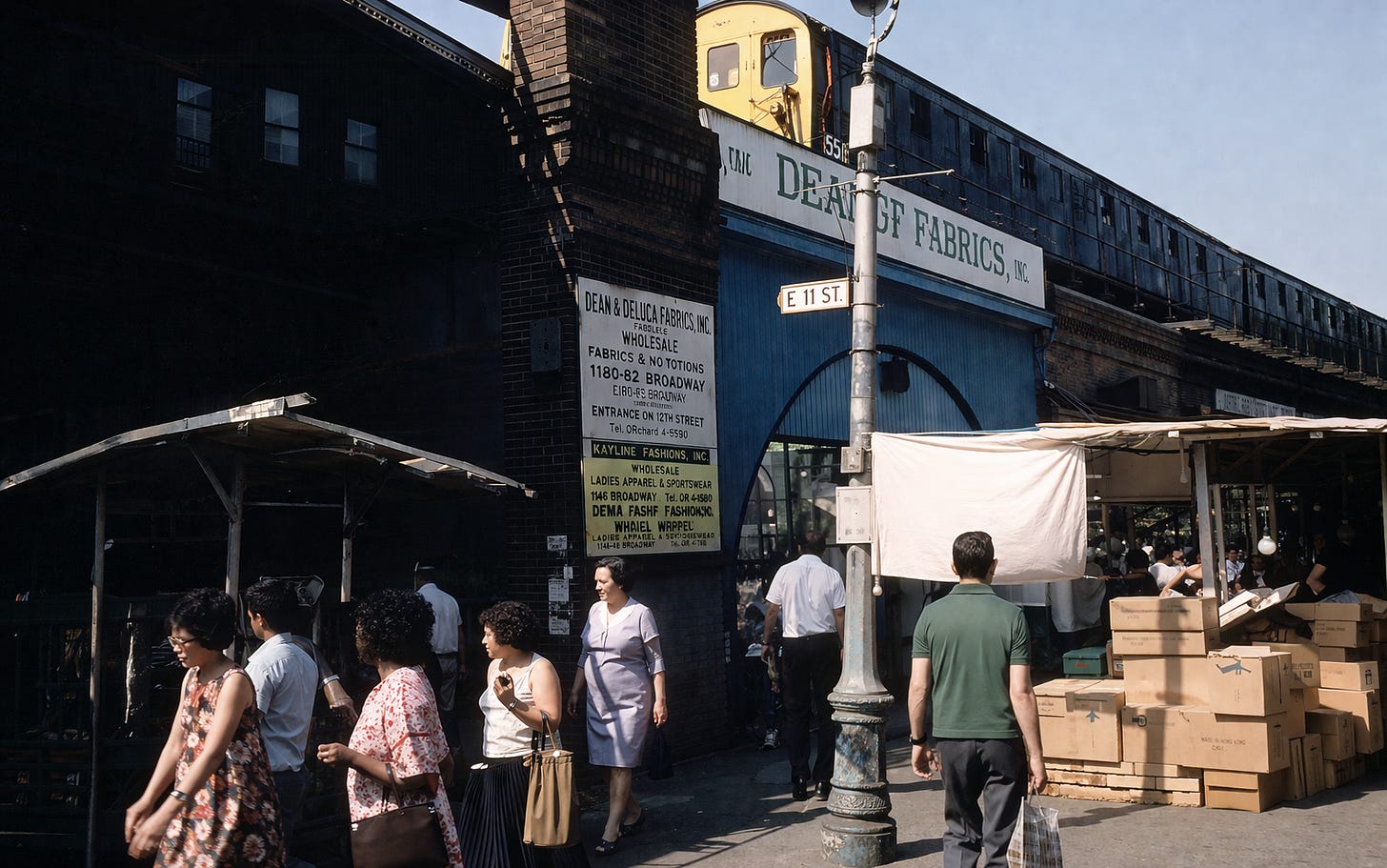 A busy street market unfolds beneath an elevated rail line, where migrant traders and small wholesalers helped shape the fabric trade that powered inner-city economies in 1970s London.