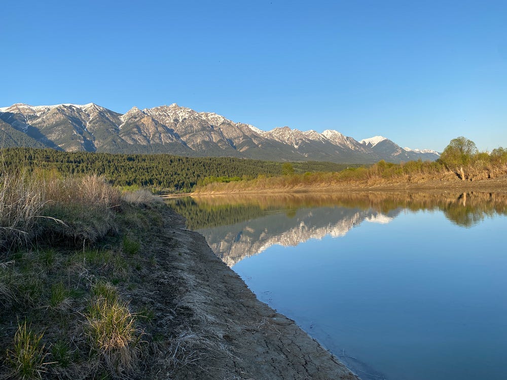 Snow-capped mountains loom in the distance reflecting in a still body of water.