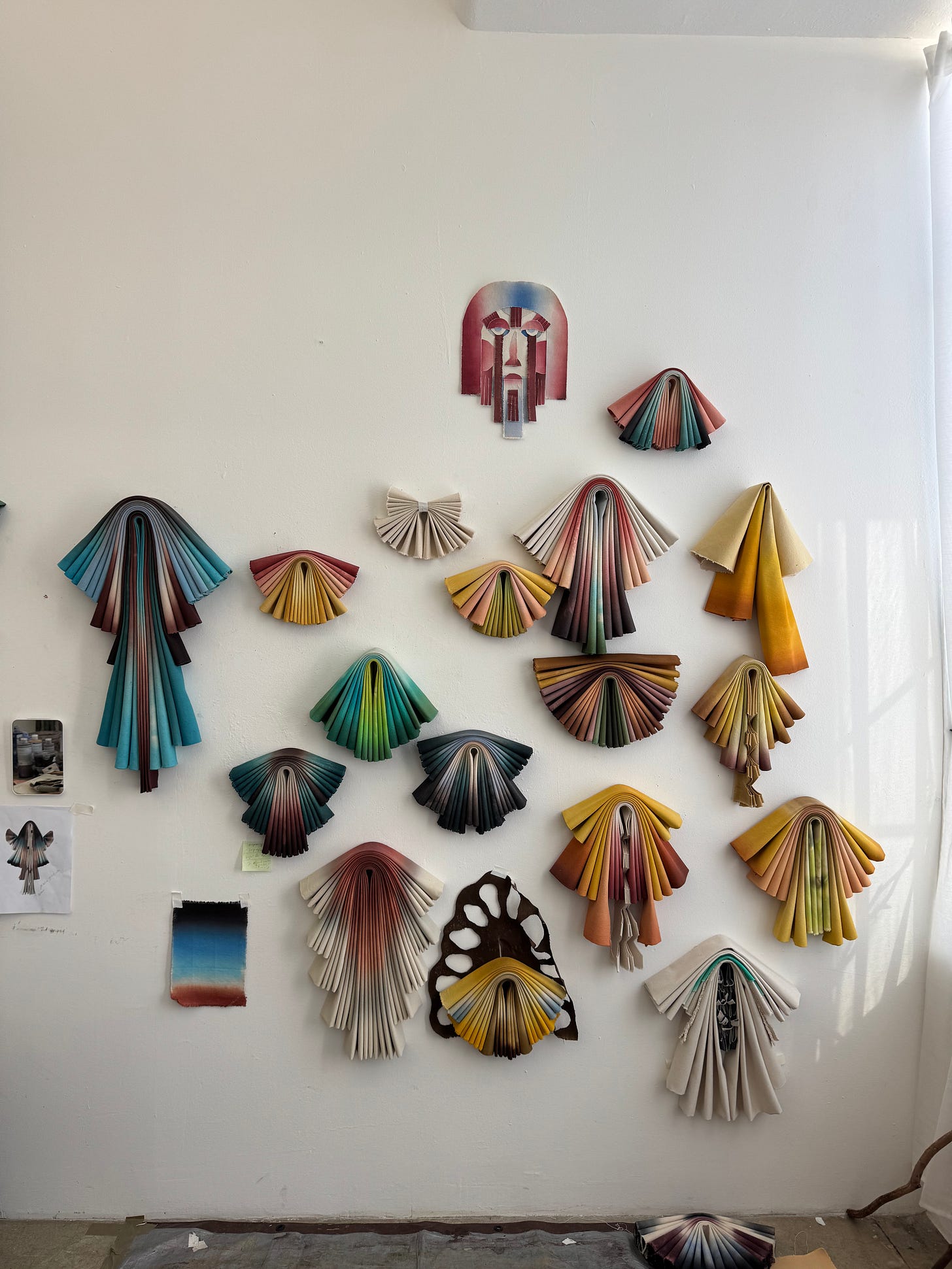 Studio wall with colorful folded-paper fan artworks arranged in a grid; photographed in daylight.