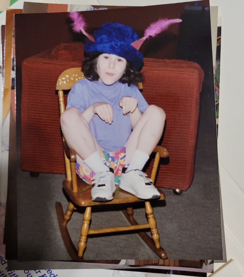 Young Lyric, in a rocking chair, crouched like a bunny, with a blue hat and pink feather ears. 