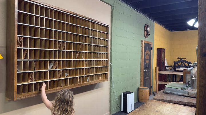 A curly-haired girl picking out cans of vegetables from an interactive mercantile. | Close up of black, lace-up ladies boots. | Low angle from behind the sales counter showing off several cupboards and an old cash register. | Same girl checking mail in the postal cubbies.