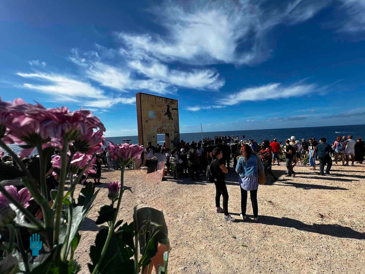 Crowd gathered at the Porta d’Europa memorial in Lampedusa overlooking the Mediterranean Sea, with flowers in the foreground and the monument facing the water.