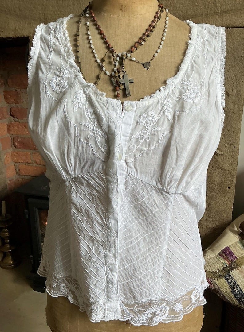 May include: A white sleeveless blouse featuring a button-down front, delicate floral embroidery on the chest, and diagonal stitched detailing across the bodice. The garment has a frayed neckline and a sheer lace trim along the bottom hem.