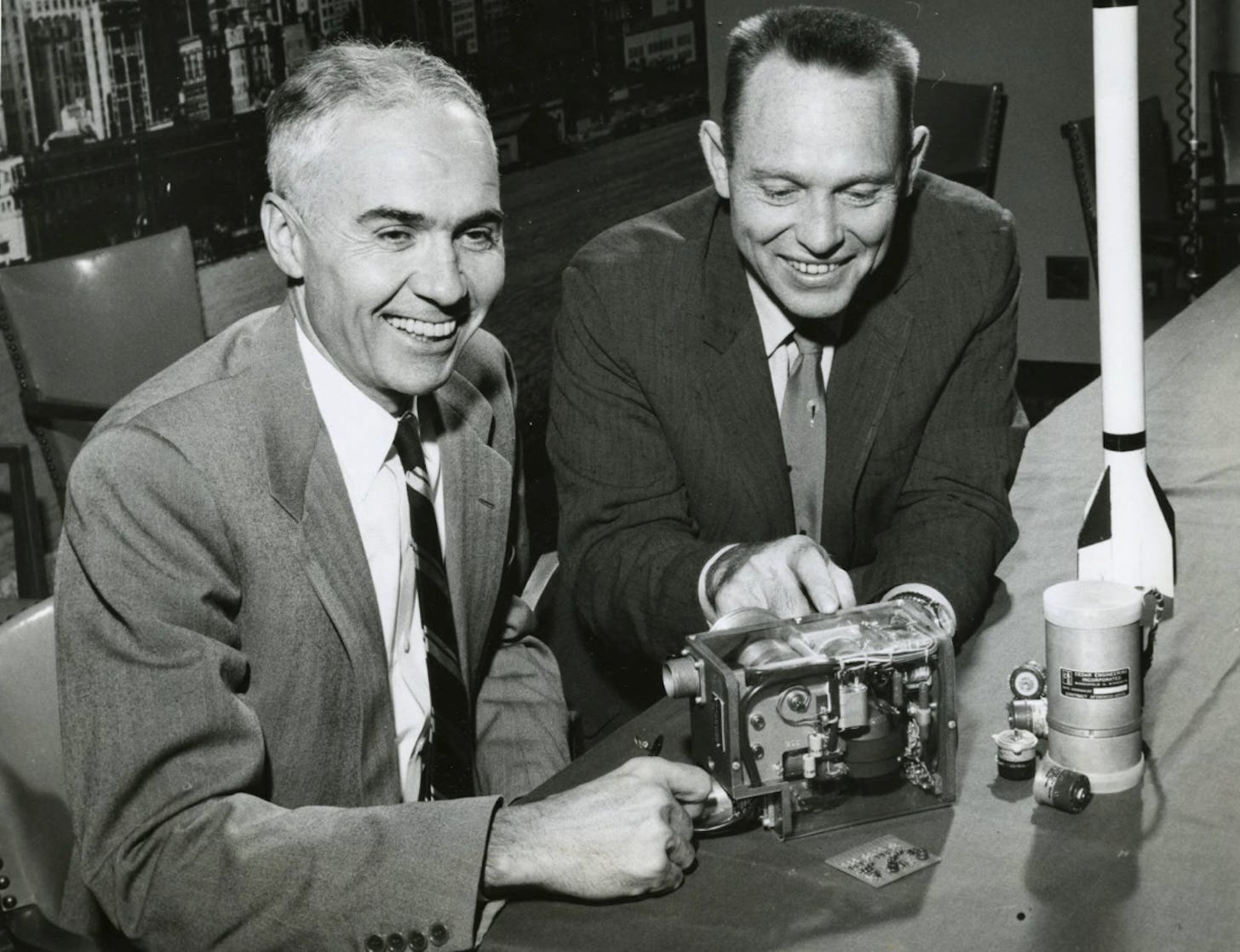 Black-and-white photograph of two men in mid-century business suits seated at a table examining a small electronic device. Bill Norris, founder of Control Data Corporation, on the left, with E. J. Manning, president of Cedar Engineering, on the right. A model rocket and additional electronic components sit on the table. Photographed November 1957 the day Control Data announced the Cedar acquisition.