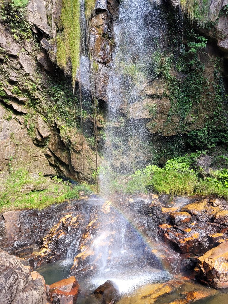 Cachoeira alta vista de frente, nem dá pra ver o topo. Ela cai numa região pedregosa com pequena área para banho e a iluminação faz surgir um pequeno arco-íris na altura das pedras.