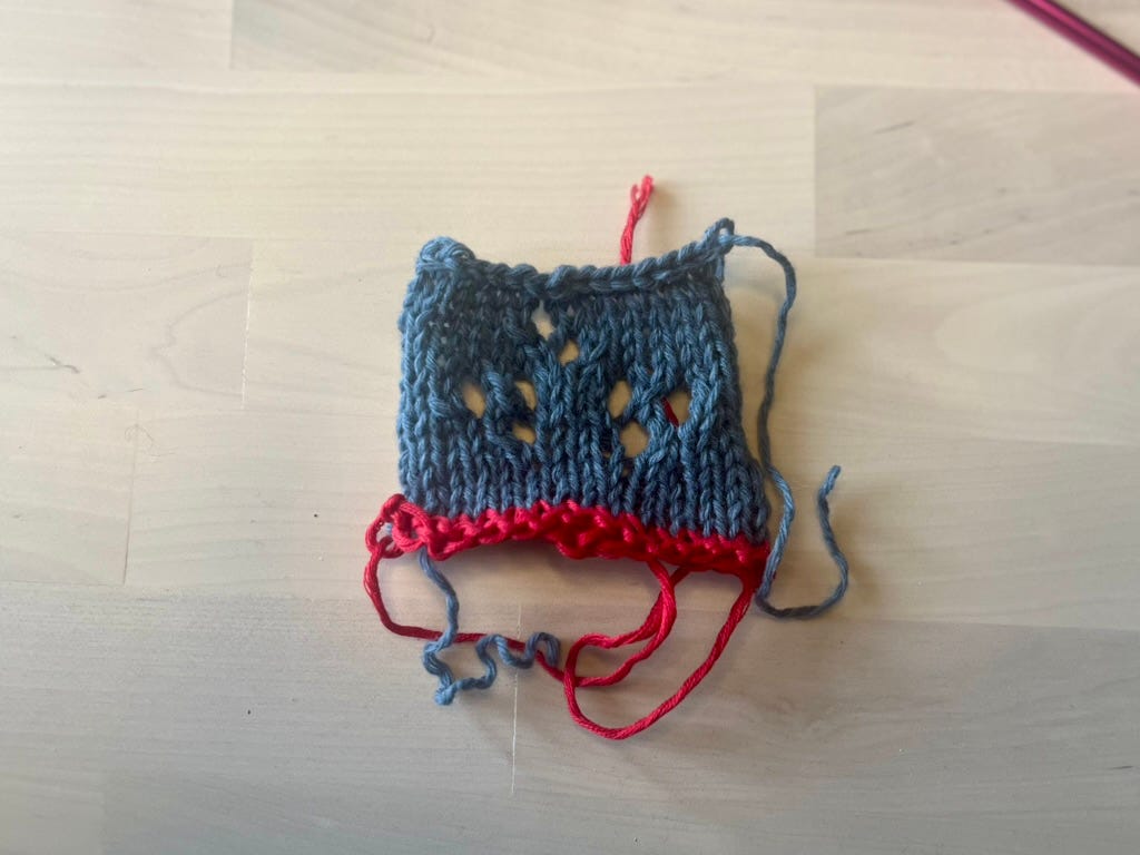 A small knitted swatch lying flat on a wooden surface showing blue-grey lace above a band of red waste yarn, with loose yarn ends visible.