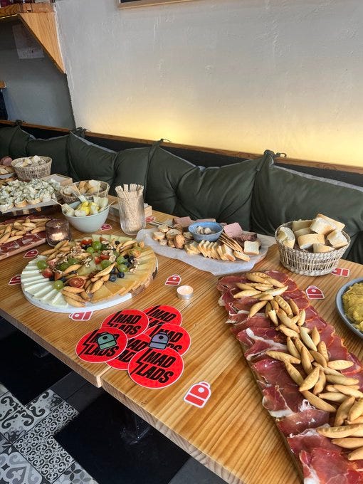 A wooden table with various platters of food, including sliced meats, breadsticks, and tapas. Bowls of olives and other appetizers are visible. Green cushioned benches line the walls, and the floor has a black and white tiled pattern. Red circular stickers with text reading "We Are One" and "Lupo" are on the table.