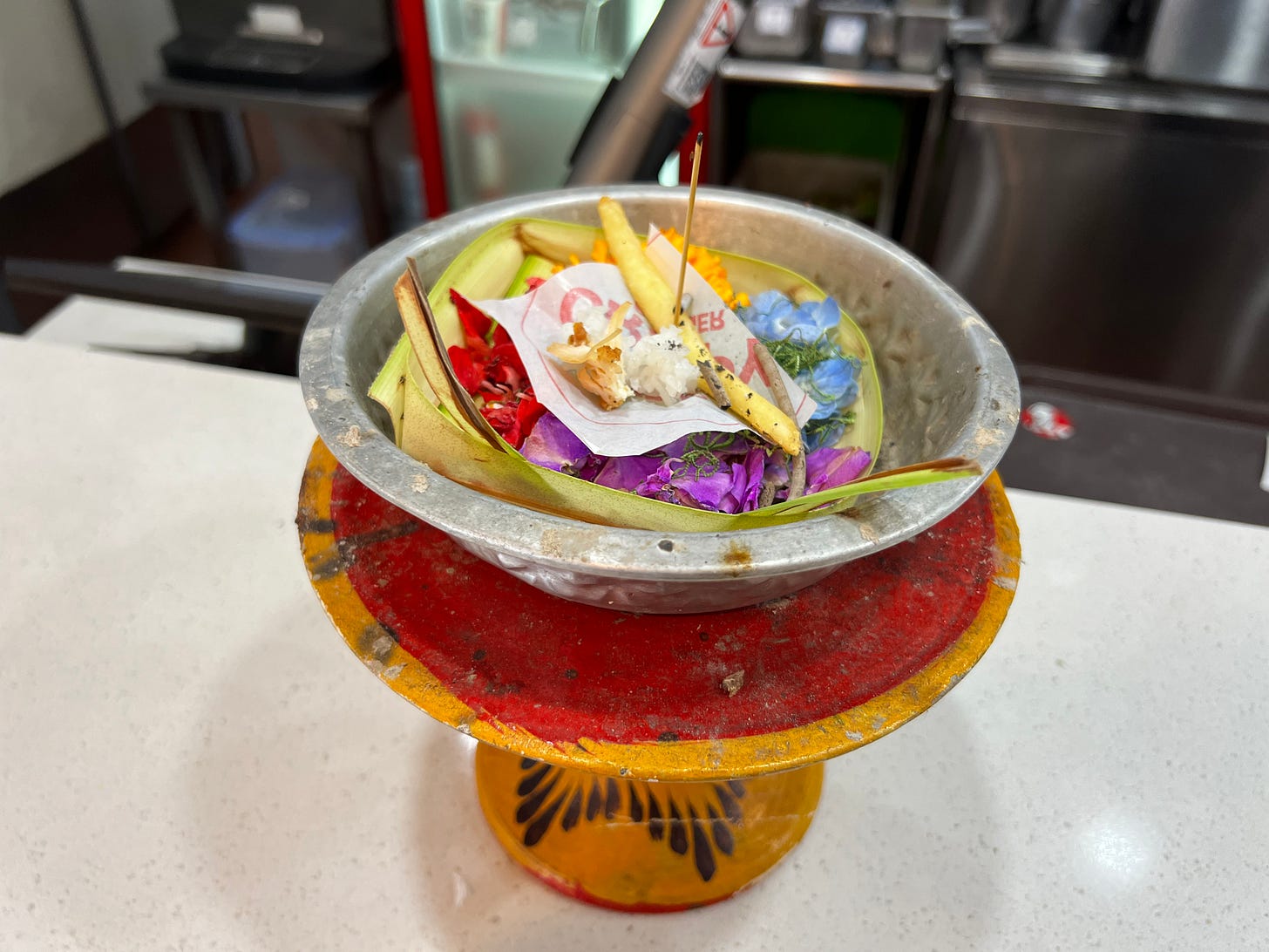 A small offering on the counter: a square of banana leaf holding flowers, a few french fries, and a burned-down stick of incense.
