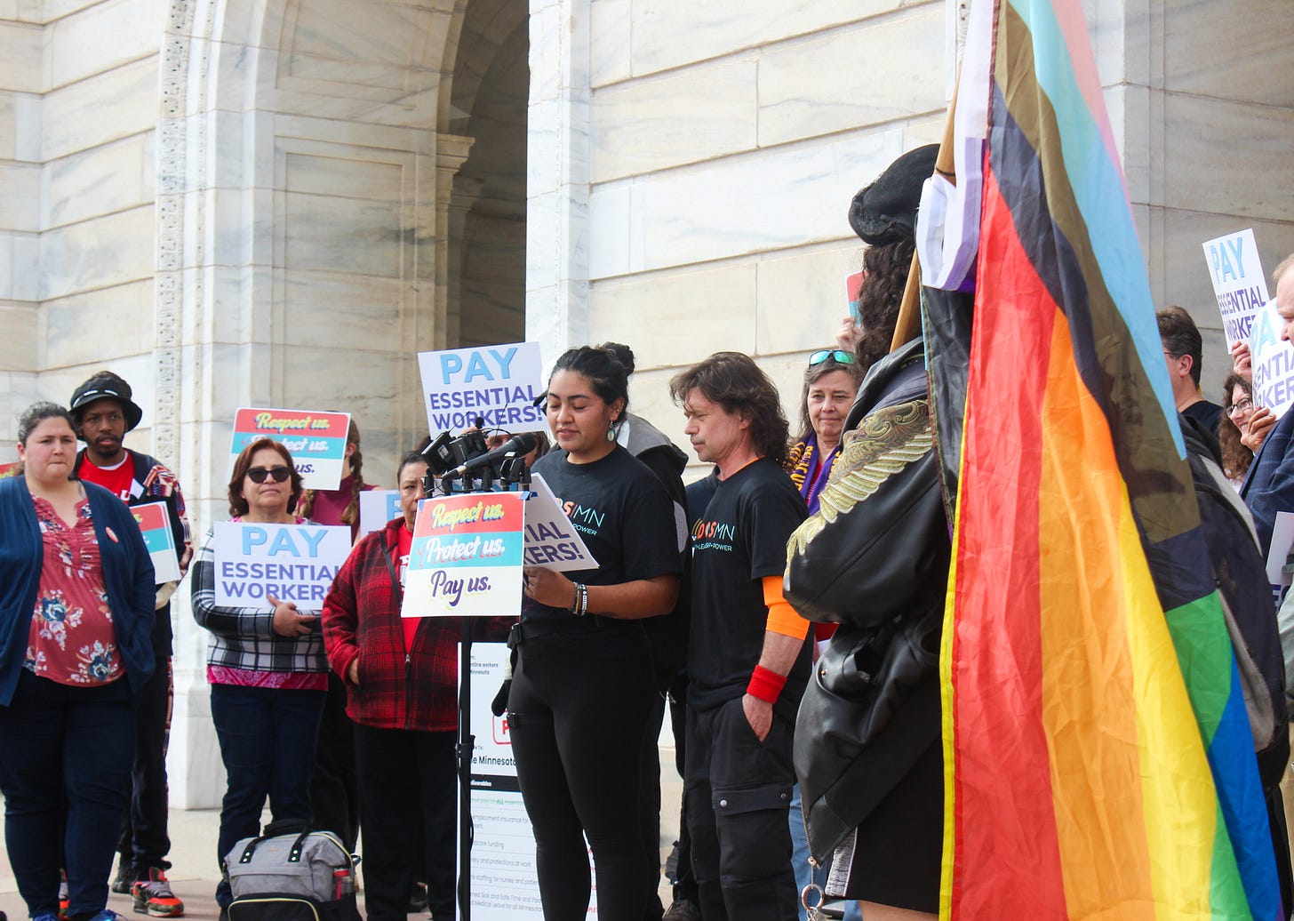 a brown woman and white man speak into microphones, surrounded by people holding "pay essential workers" signs a brown woman and white man speak into microphones, surrounded by people holding "pay essential workers" signs