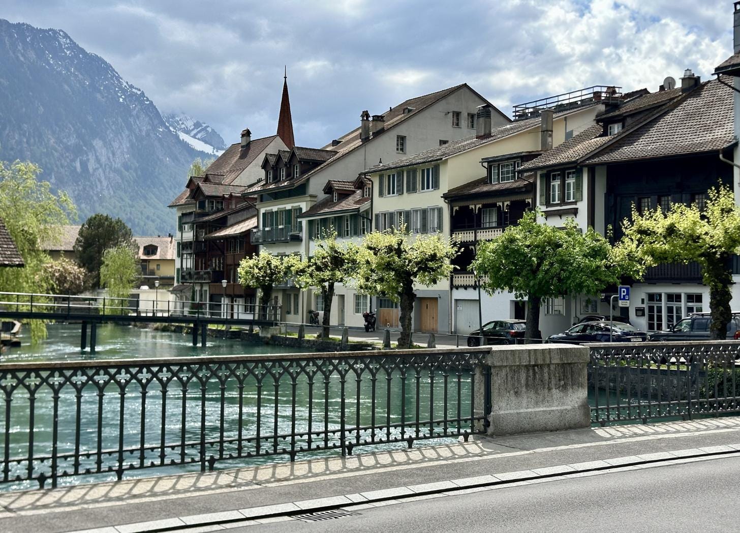 Traditional Swiss residential buildings with wooden balconies and chalet rooftops along the clear blue Aare canal in central Interlaken, with green spring trees, a church spire, and snow-capped Alpine mountains rising behind the town. Traditional Swiss residential buildings with wooden balconies and chalet rooftops along the clear blue Aare canal in central Interlaken, with green spring trees, a church spire, and snow-capped Alpine mountains rising behind the town.