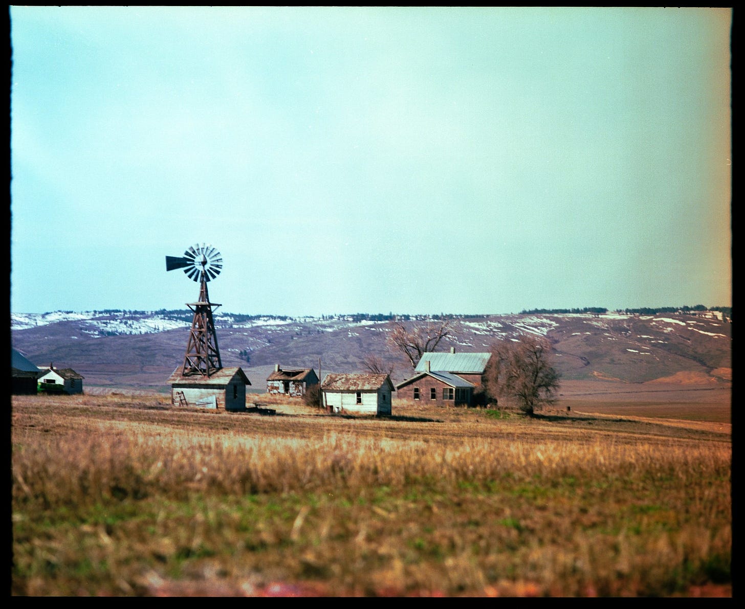 An old abandoned farmstead with various out buildings is in the middle ground. Snow-covered hills rest beyond. One of the buildings has a windmill atop it. The foreground is cut, dry grass. 