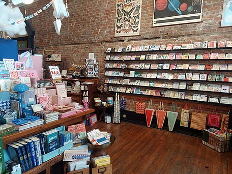 A photograph of a stationery store with brick walls lined with shelves of cards, books, and fancy paper. A photograph of a stationery store with brick walls lined with shelves of cards, books, and fancy paper.