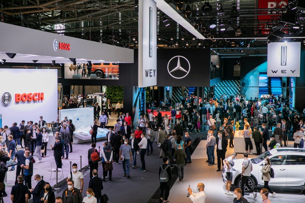 Una foto realista de un gran salón de exposiciones con varias marcas de coches que muestran sus vehículos. La imagen está tomada desde una posición elevada, mirando hacia el suelo de la exposición. El salón está iluminado con múltiples focos. Las marcas visibles en la imagen son Bosch, Mercedes-Benz y WEY. El stand de Bosch tiene una gran pancarta blanca con el logo de la empresa en rojo y negro. El stand de Mercedes-Benz tiene una gran pancarta negra con el logo de la empresa en blanco. El stand de WEY tiene una gran pancarta blanca con el logo de la empresa en negro. Hay varios coches en exhibición en cada stand. Hay muchas personas caminando por el suelo de la exposición, mirando los coches en exhibición.