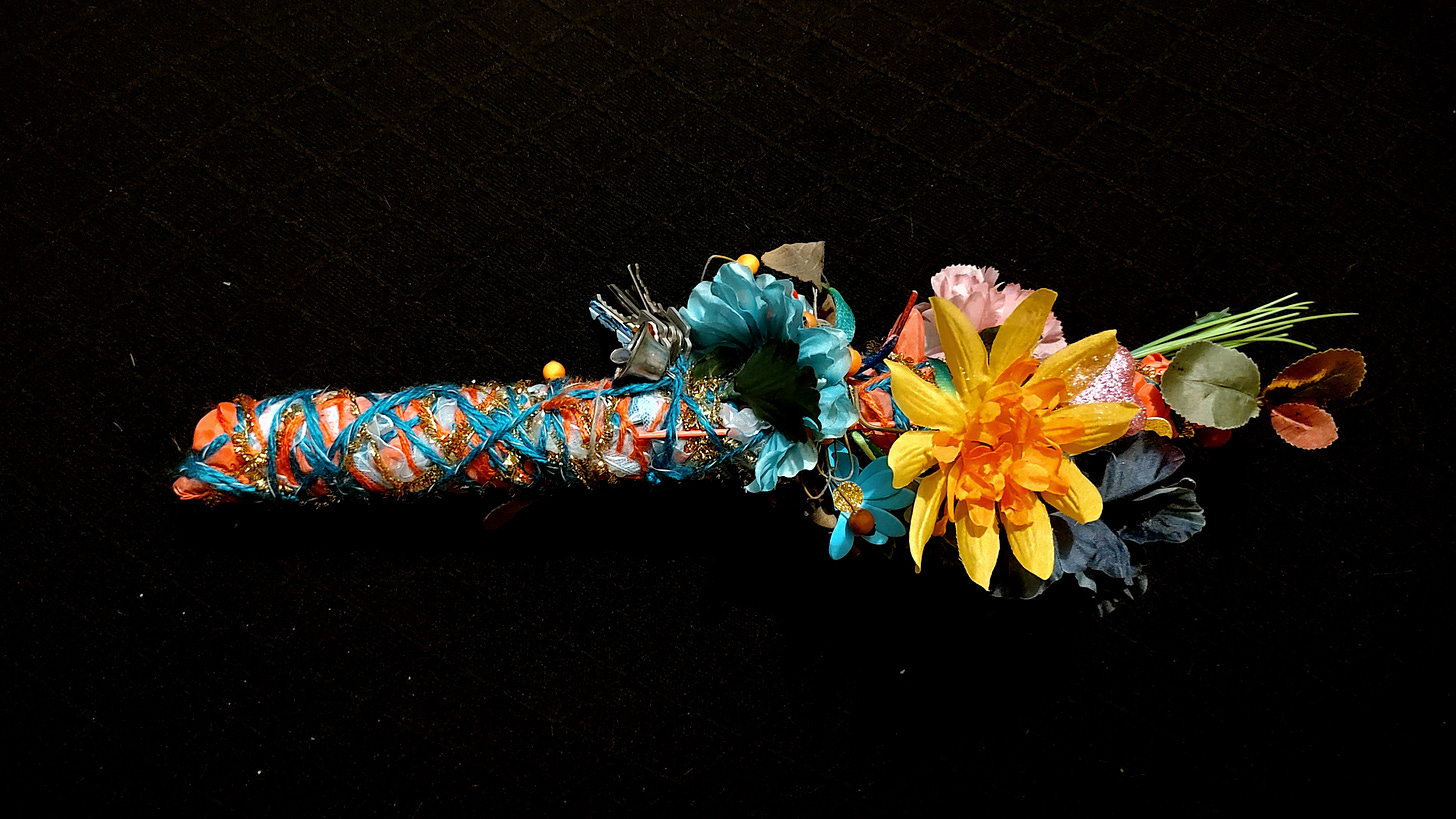 A wand wrapped in colorful textiles, with silk flowers, keys, a bell and sparkly objects.
