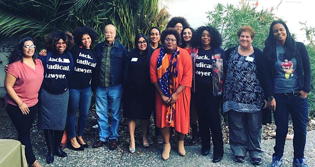 Peggy stands in a group of 12 people, mostly Black women. Several (including Peggy) wear a black T-shirt with white text that reads: Black. Radical. Lawyer."