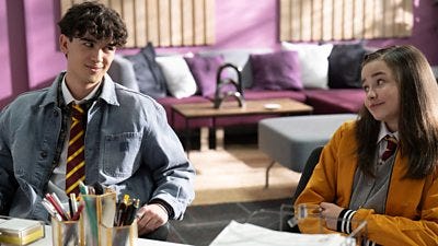 A boy and girl in school uniform sit at the head teacher's desk, smirking to each other