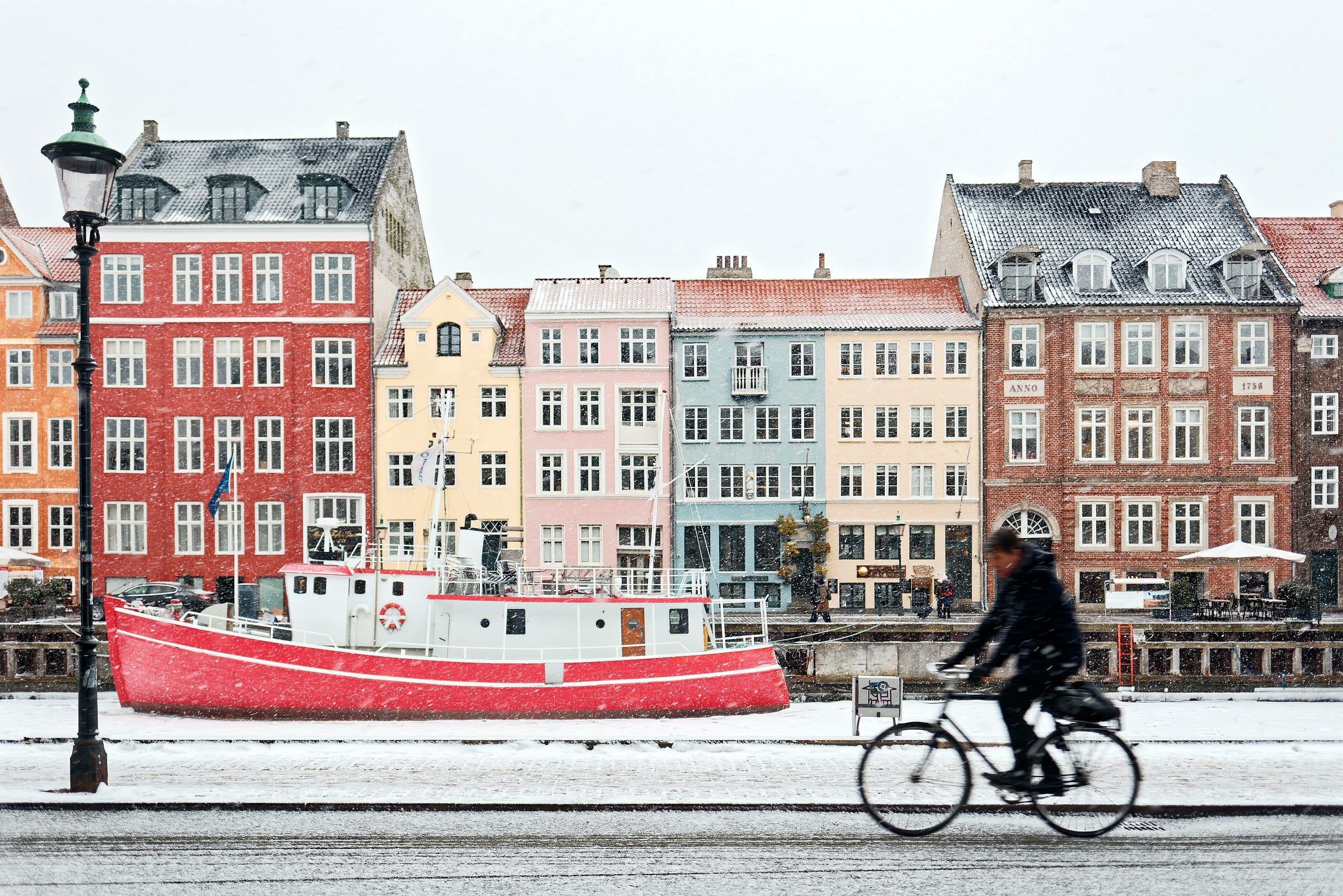 A scene from Copenhagen, showing some houses in Nyhavn from across the canal — a boat is in the canal and a bicyclist passing in front of the camera. It is winter and small amount of snow is coloring the roofs slightly white. A scene from Copenhagen, showing some houses in Nyhavn from across the canal — a boat is in the canal and a bicyclist passing in front of the camera. It is winter and small amount of snow is coloring the roofs slightly white.