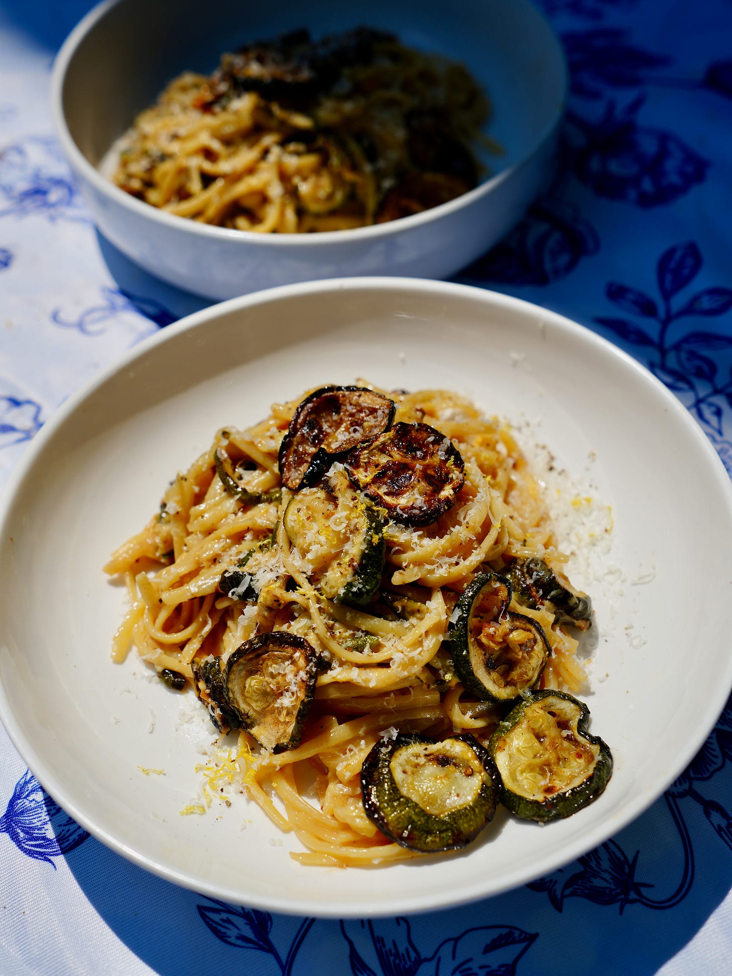 A bowl of creamy miso Alfredo linguine topped with caramelized zucchini and grated Parmesan on a blue floral tablecloth.