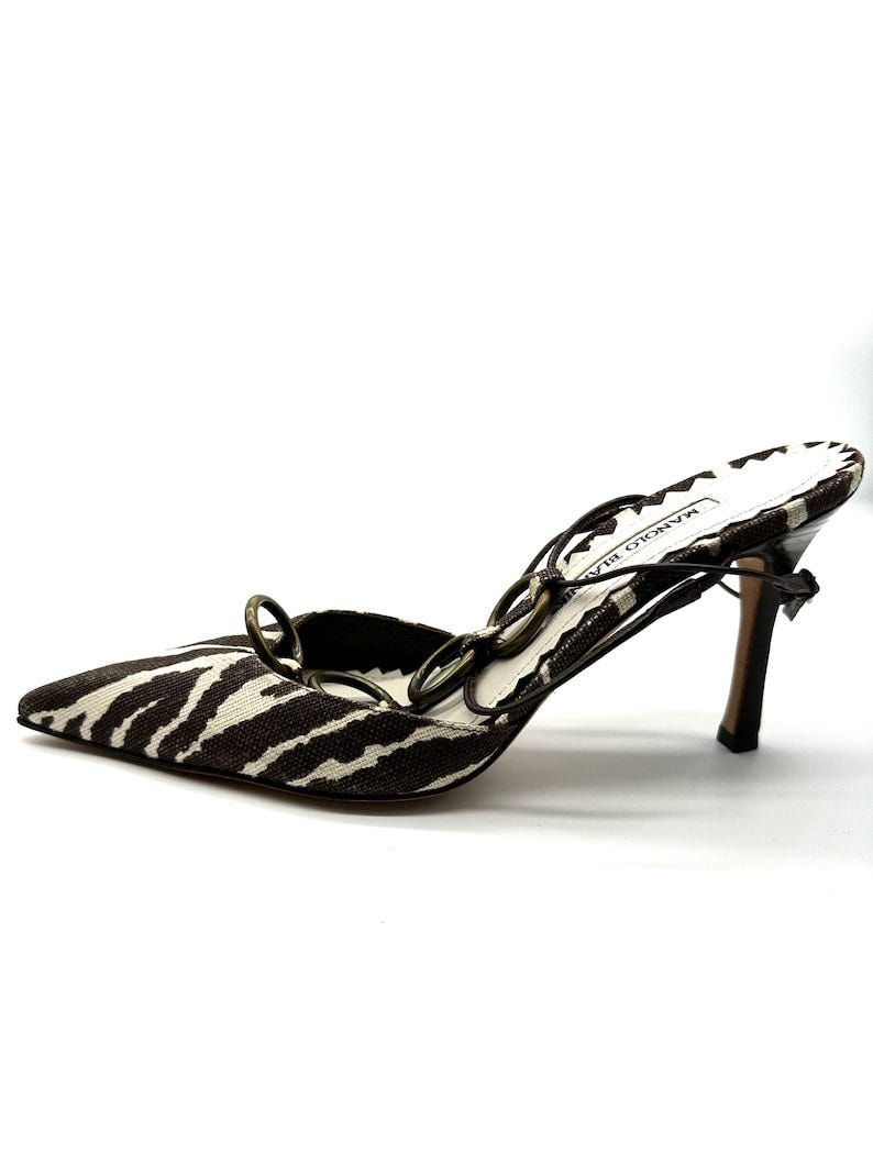 May include: Zebra print high heel shoes with a black and white pattern and a gold chain detail. The shoes are labeled 'MANOLO BLAHNIK'.