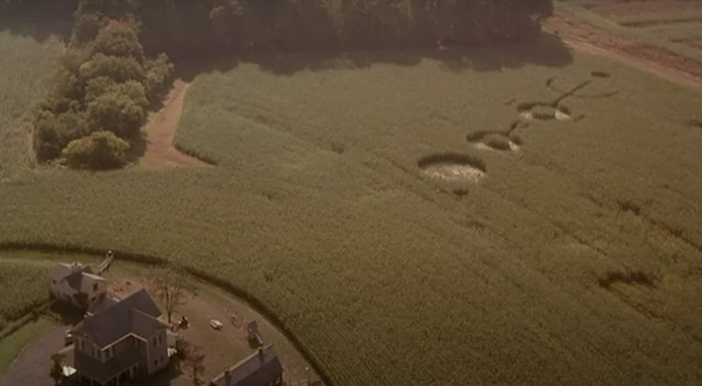 An aerial view of green crops with a farmhouse at the bottom left, and three large crop circles done by aliens.