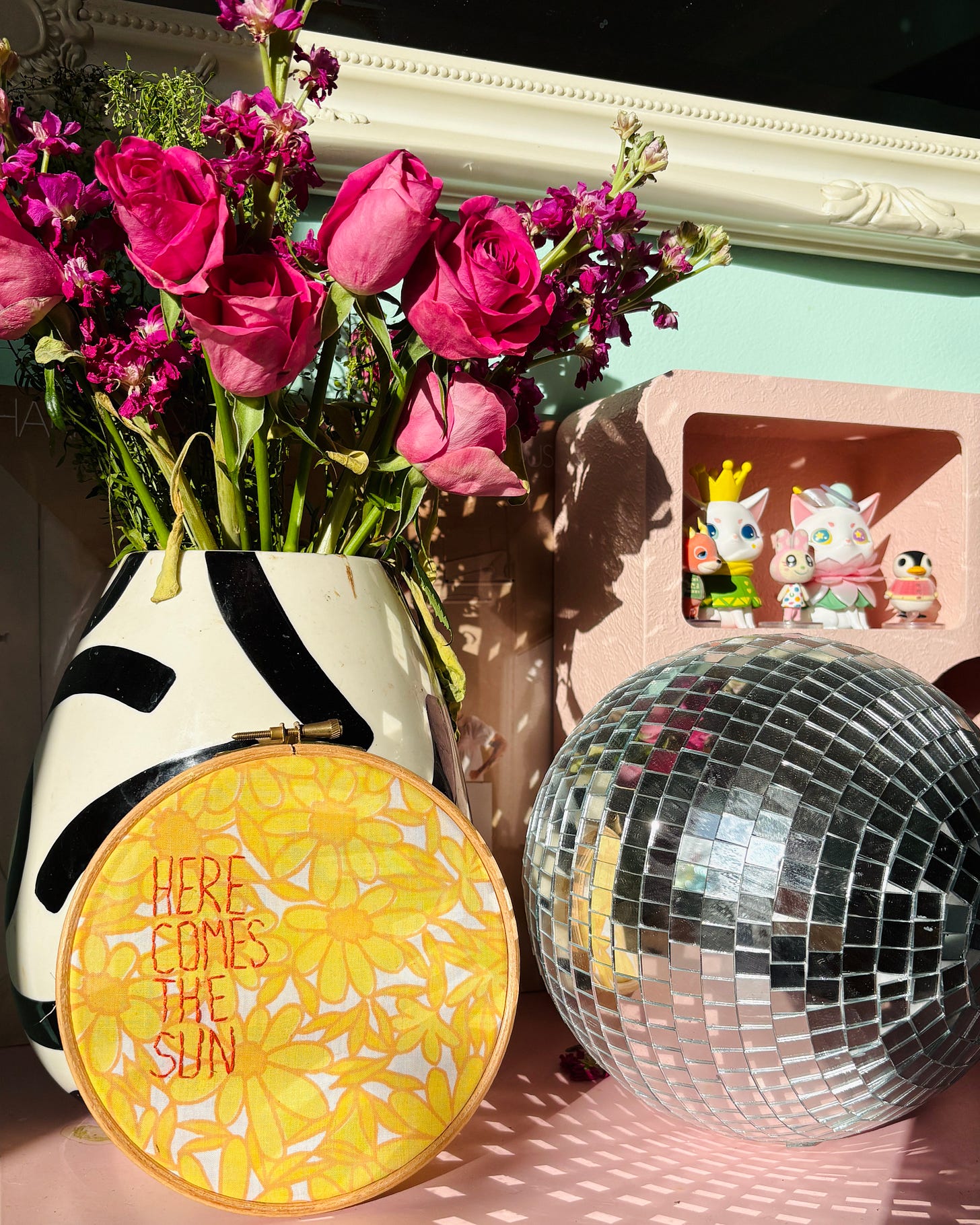 A photo of a black and white vase filled with pink roses and stocks next to a disco ball on a pink unit with the sun shinning on it