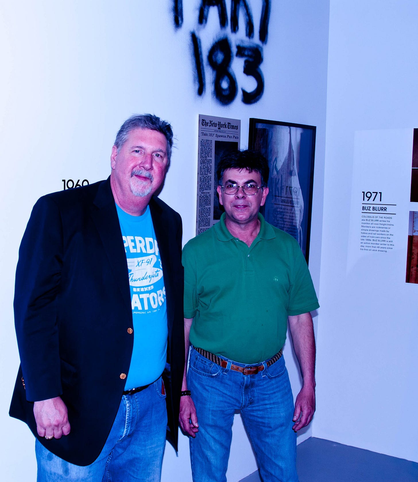 Two men posing in a gallery. On the right, TAKI 183 in a green polo shirt, under his name sprayed on the wall.