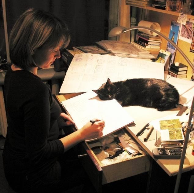 This may contain: a woman sitting at a desk with a cat laying on top of her book and papers