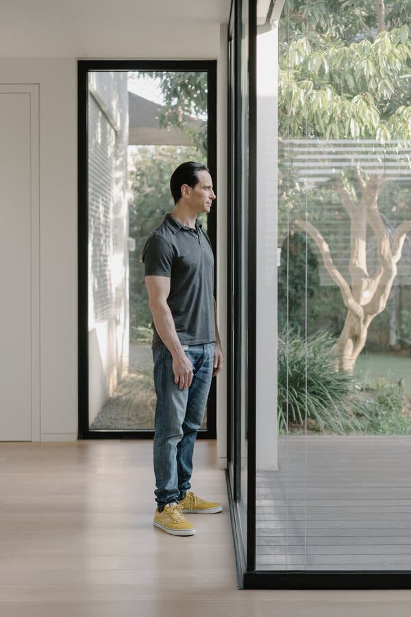 Adam Fisher, in a dark polo shirt, jean and yellow sneakers, standing by floor-to-ceiling windows and looking out at outdoor greenery and a tree. Adam Fisher, in a dark polo shirt, jean and yellow sneakers, standing by floor-to-ceiling windows and looking out at outdoor greenery and a tree.
