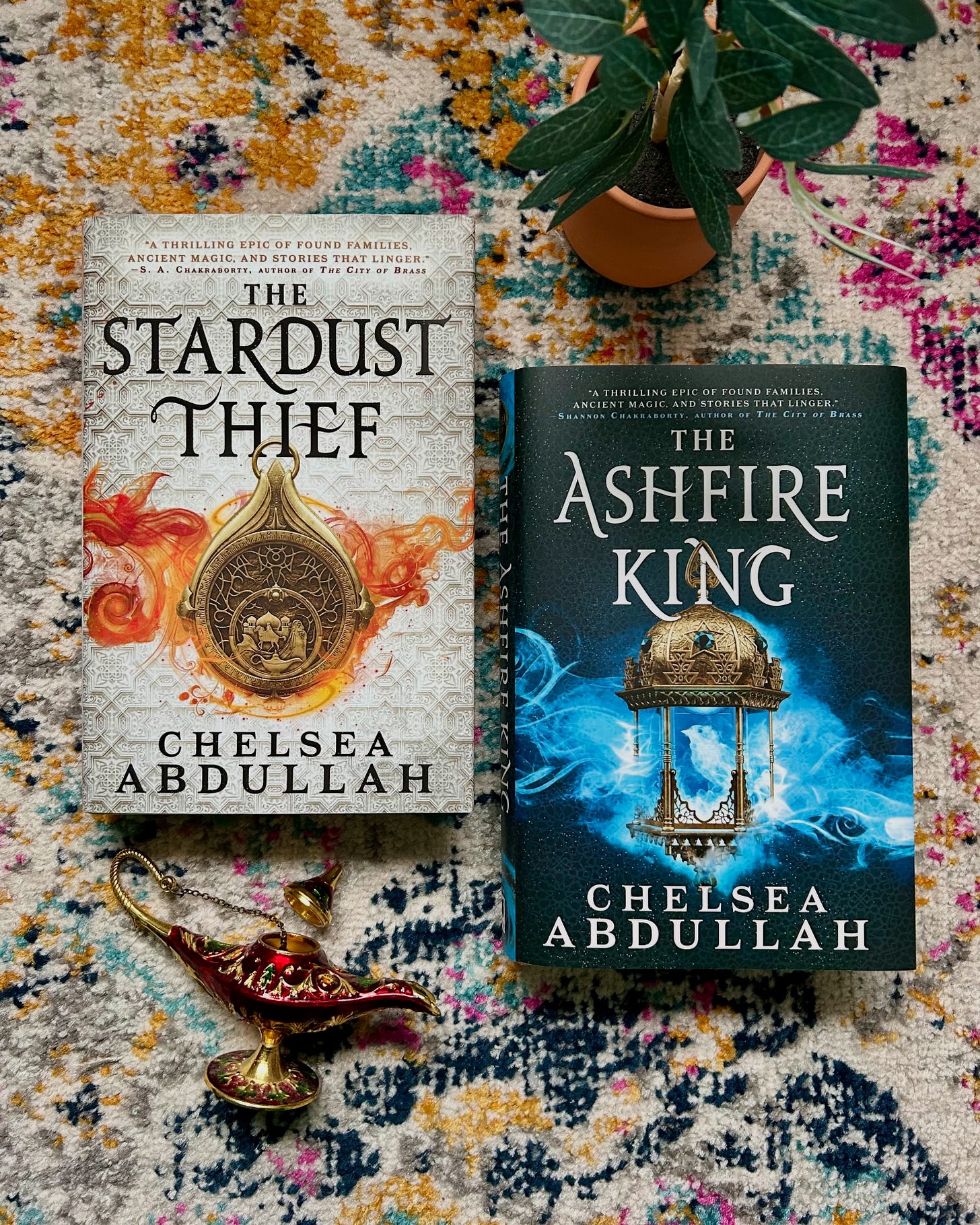 THE STARDUST THIEF hardcover and THE ASHFIRE KING hardcover lie side by side on a patterned rug. An open oil lamp lays beneath them.