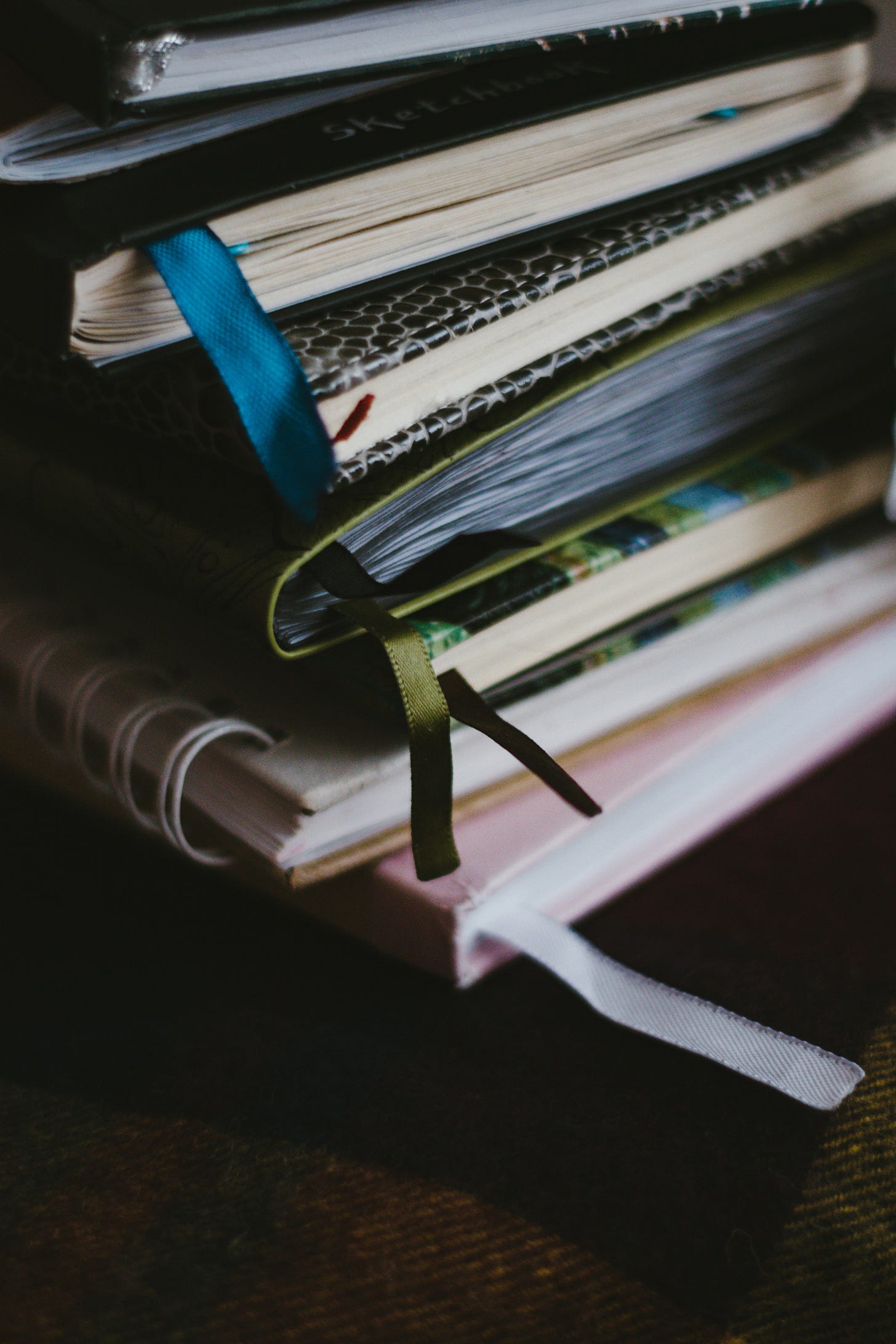 A close-up of a stack of colorful, well-worn journals and sketchbooks with fabric bookmarks sticking out. The pages appear slightly bent, showing signs of frequent use. The journals rest on a soft, dark surface, evoking a feeling of intimacy and reflection. A close-up of a stack of colorful, well-worn journals and sketchbooks with fabric bookmarks sticking out. The pages appear slightly bent, showing signs of frequent use. The journals rest on a soft, dark surface, evoking a feeling of intimacy and reflection.