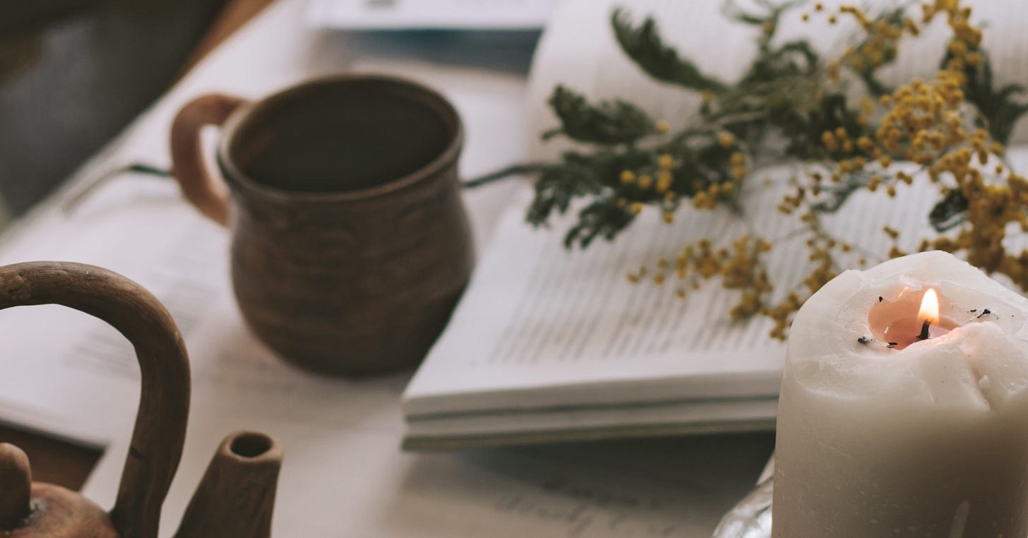 A cosy workspace with a candle, a pottery mug, fresh greenery, and scattered books and papers on a wooden table.
