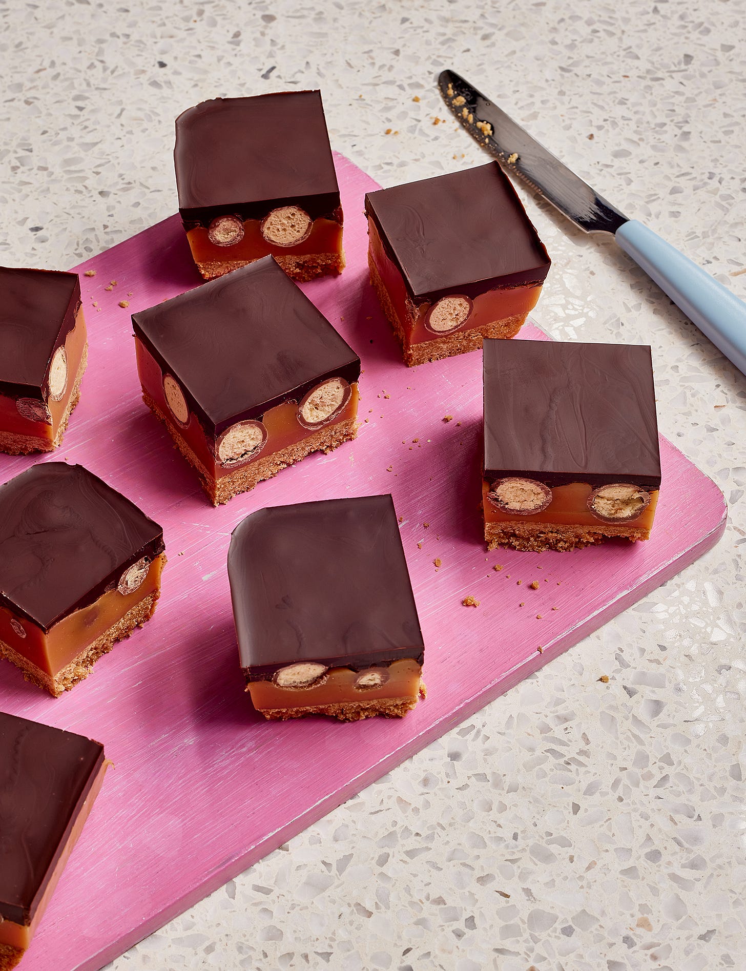 Sqaures of chocolate caramel shortbread with maltesers on a pink chopping board