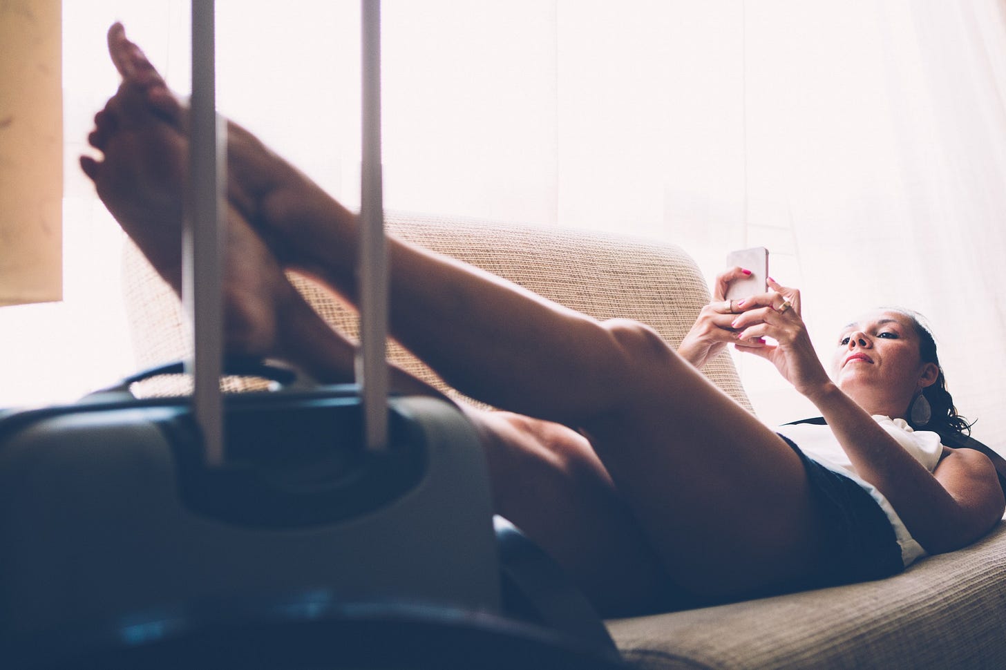 A woman stares blankly at her phone. Her feet are propped up on a suitcase.