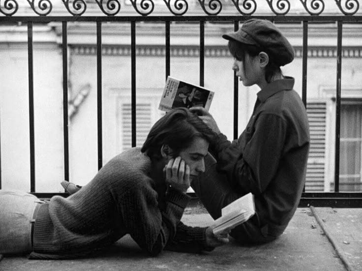 This may contain: two people sitting on the ground reading books and talking on their cell phones in front of a fence This may contain: two people sitting on the ground reading books and talking on their cell phones in front of a fence